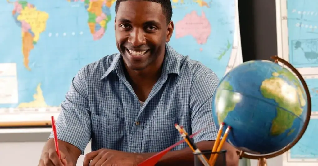 Smiling teacher sitting at his desk, after thinking positive affirmations for teachers about work load.