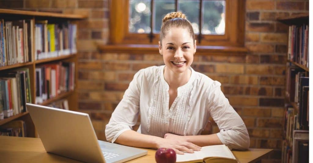 Smiley teacher sitting at a desk with a laptop with rows of books behind her thinking positive affirmations for teachers.