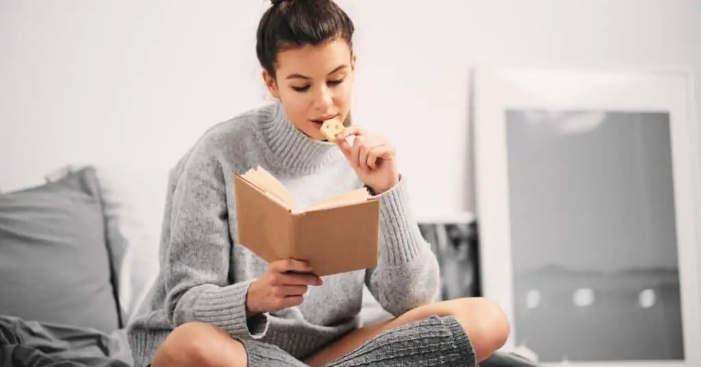 Woman reading a book on the bed  while she eats a cracker.